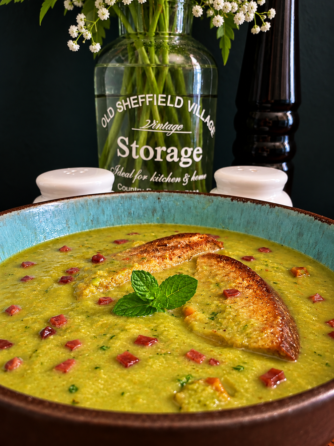Creamy English pea and mint soup in a rustic turquoise ceramic bowl, garnished with crispy vegan bacon cubes, fresh mint leaves and two slices of golden lemon farmhouse bread, photographed against a dark green kitchen background with a vintage glass vase.