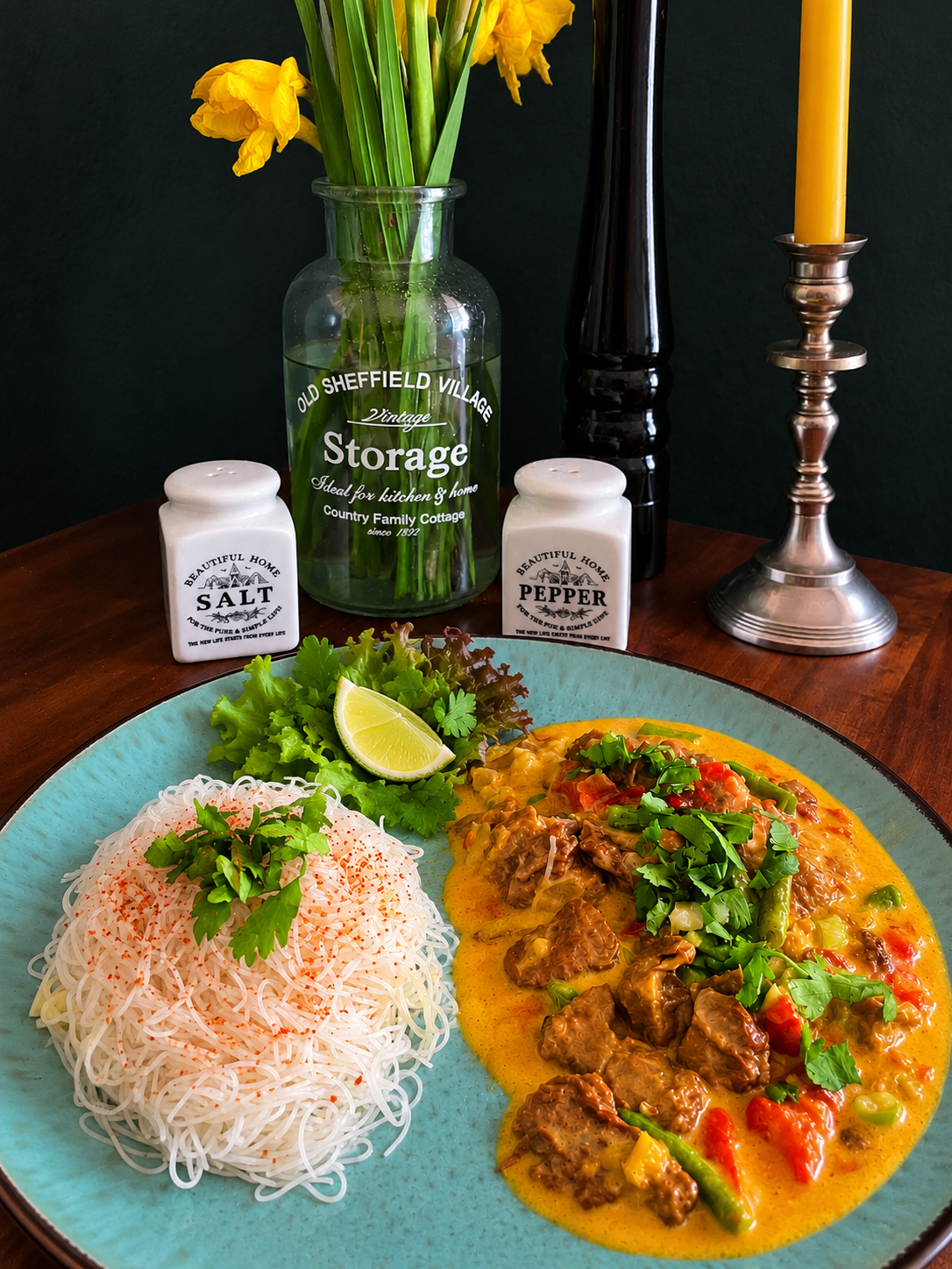 2. IMAGE ALT TEXT Vegan Lao-inspired lemongrass and coconut curry with Mock Duck, green beans, tomatoes and fresh coriander, served with thin rice noodles sprinkled with chili on a turquoise plate, garnished with lettuce and lime.