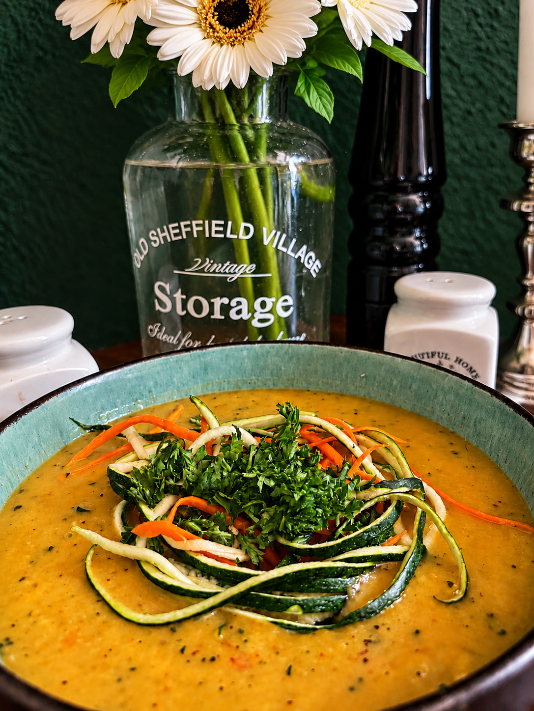 Creamy vegan Serbian zucchini and carrot soup in a turquoise ceramic bowl, topped with fresh zucchini and carrot ribbons and chopped parsley, served against a dark green background.
