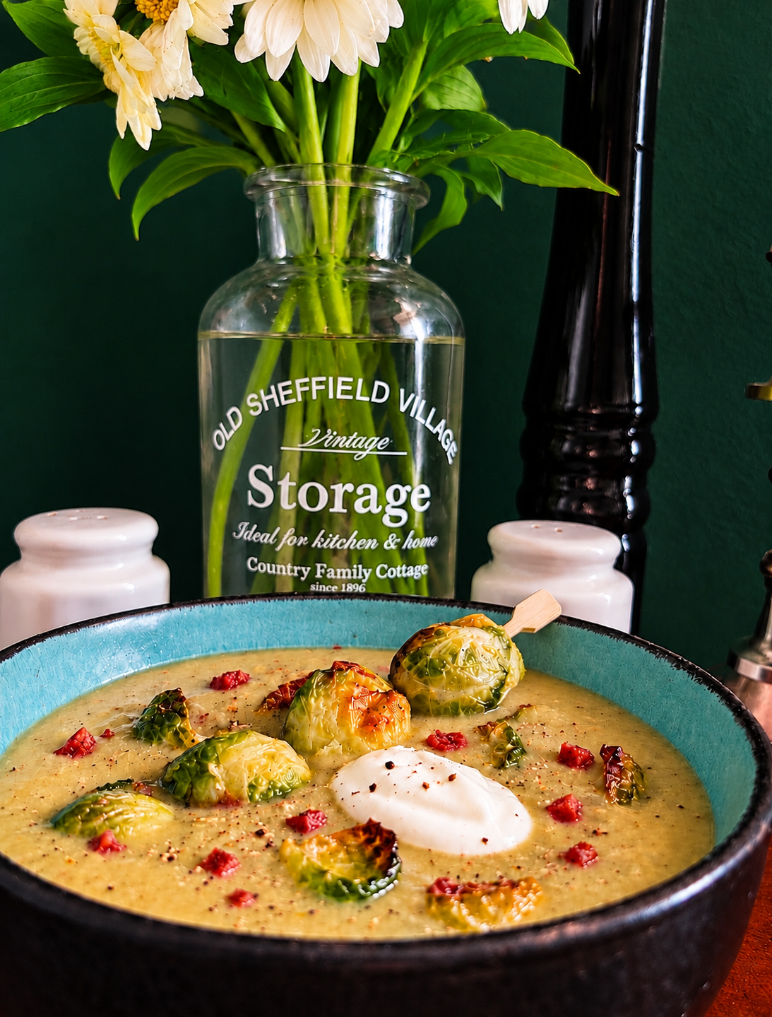 Creamy Belgian Brussels sprout soup in a turquoise bowl, garnished with roasted Brussels sprouts, crispy vegan ham cubes, vegan sour cream and a maple-glazed sprout skewer.