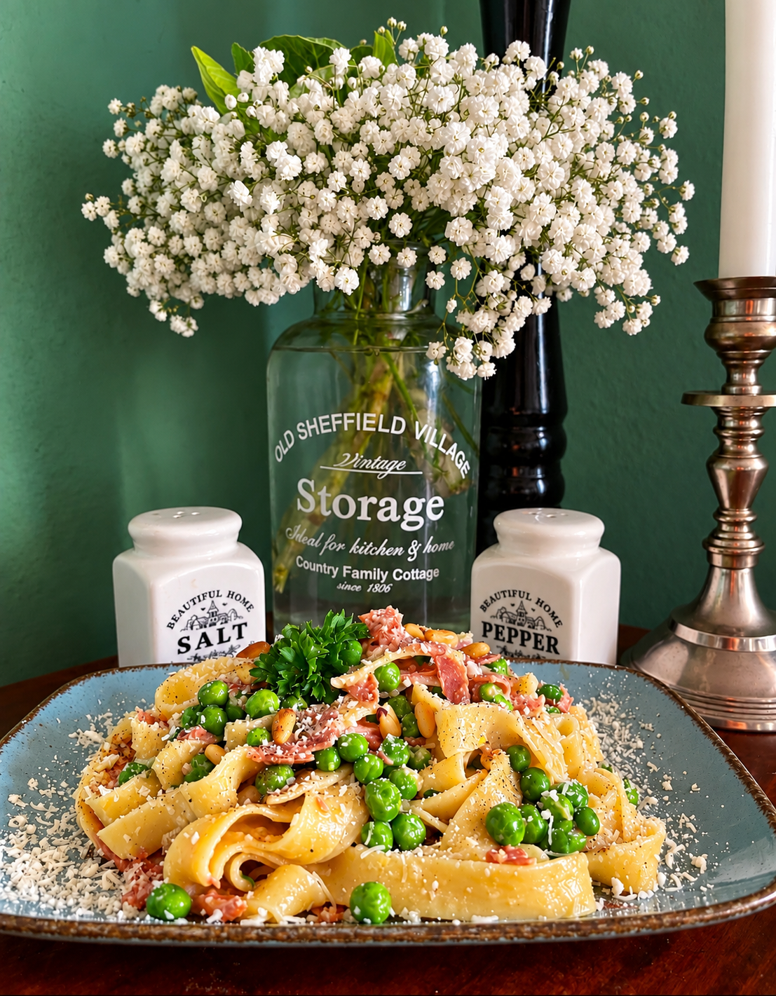 Vegan Fettuccine alla Papalina with bright green peas, plant-based ham strips, grated vegan Parmesan, toasted pine nuts and fresh parsley, served on a blue-grey ceramic plate with white flowers in the background.