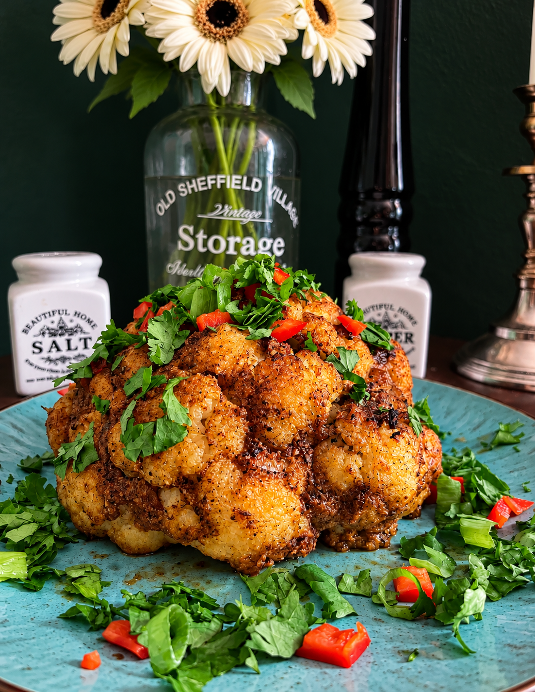 Whole Roasted Jerk Cauliflower with a deeply golden-brown spiced crust, topped with fresh coriander and chopped red chili, served with lime on a rustic turquoise ceramic plate.